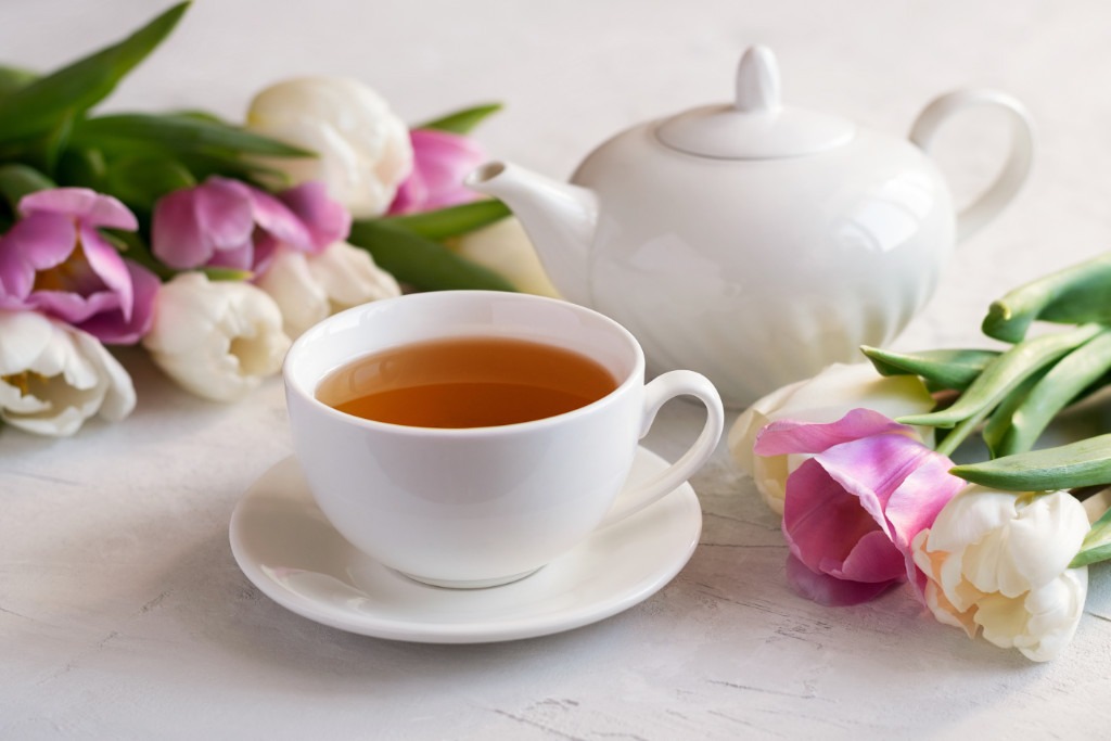 Fragrant tea in a white cup with a teapot and a bouquet of fresh tulips on a bright table. Close-up. Selective focus.